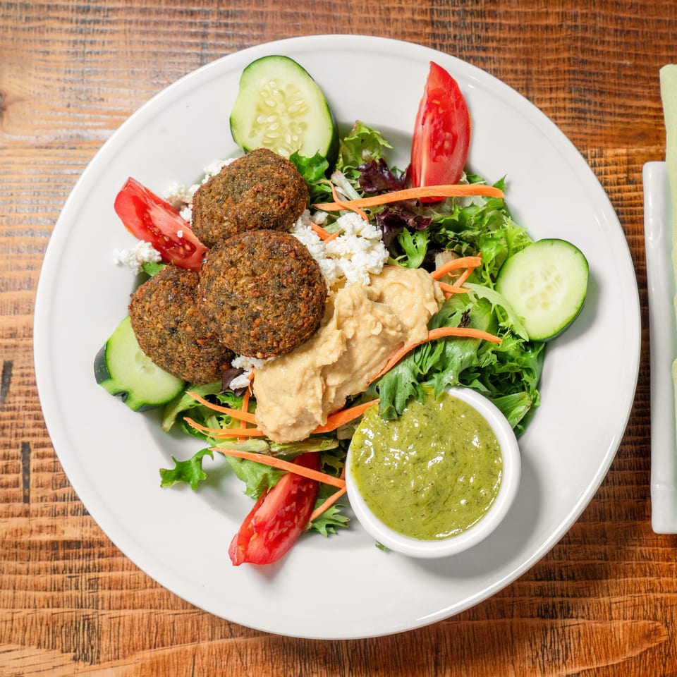 Falafel salad with hummus, feta, tomatoes, cucumbers, and green dressing on a white plate.