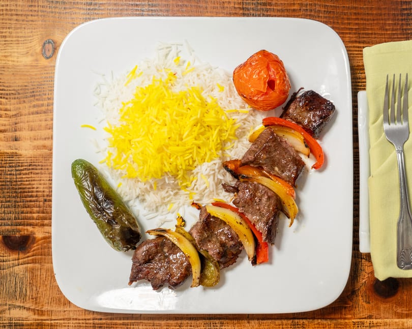 A plate of beef and vegetable skewers, rice, grilled tomato, and pepper on a wooden table.