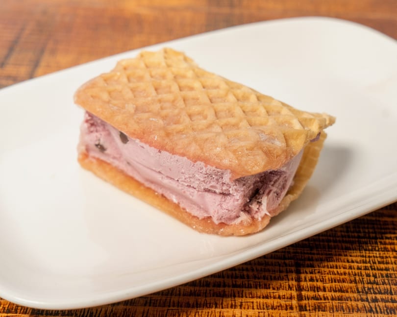 A waffle ice cream sandwich with purple ice cream and chocolate chips on a white plate.