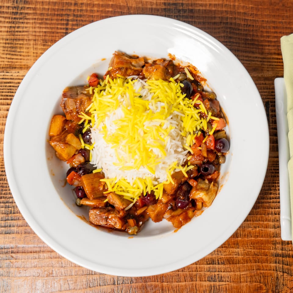 A white plate of stew with rice and yellow shredded cheese on a wooden table.