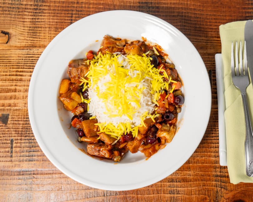 A white plate of stew with rice and yellow shredded cheese on a wooden table.