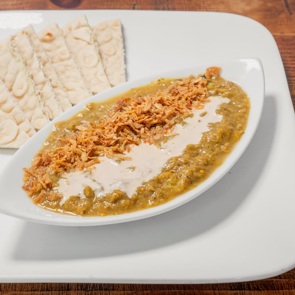 A white platter holds a boat-shaped dish of green stew with fried onions and cream, alongside flatbread.