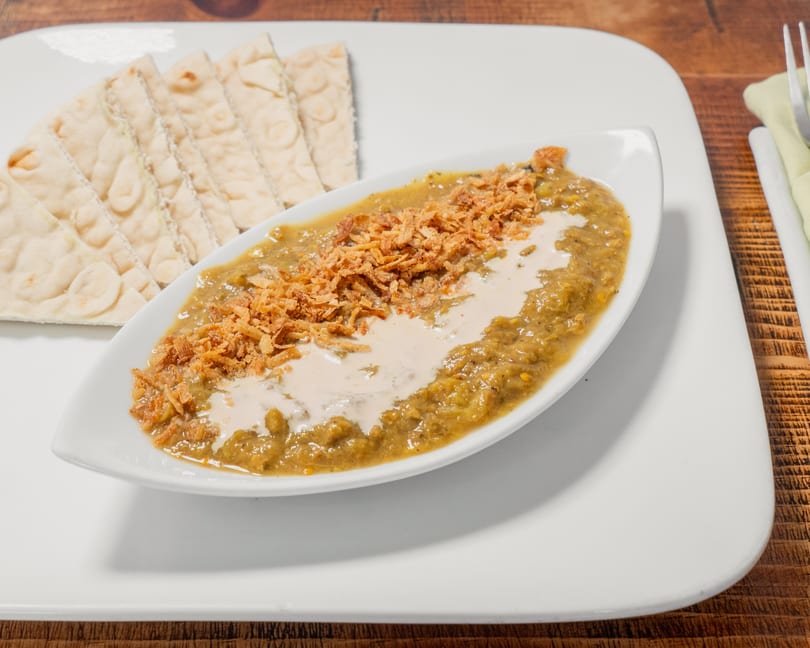 A white platter holds a boat-shaped dish of green stew with fried onions and cream, alongside flatbread.