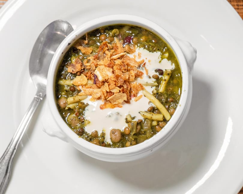 Hearty green soup with noodles, lentils, chickpeas, topped with fried onions and a creamy swirl.