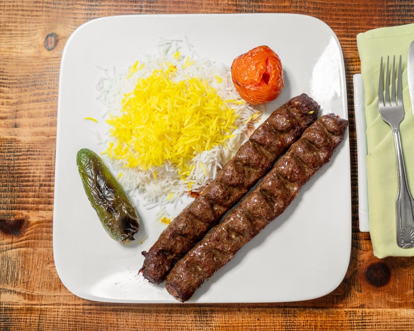 Two grilled kebabs, white rice with saffron, a roasted tomato, and a green pepper on a white plate.