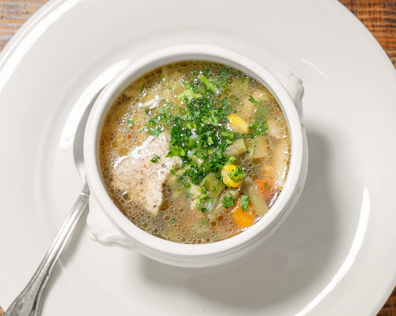 Hearty chicken and vegetable soup with parsley garnish in a white bowl on a white plate with a spoon.