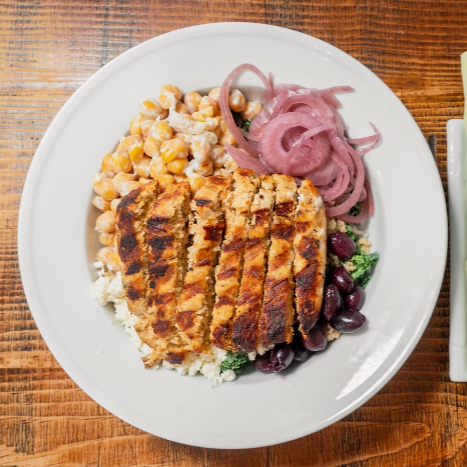 Grilled chicken, chickpeas, pickled red onions, and olives in a white bowl on a wooden table.
