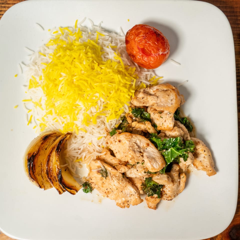 Chicken and kale with white and yellow rice, grilled onion, and a roasted tomato on a white plate.
