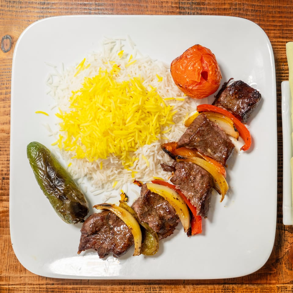 A plate of beef and vegetable skewers with white and yellow rice, a roasted tomato, and a pepper.