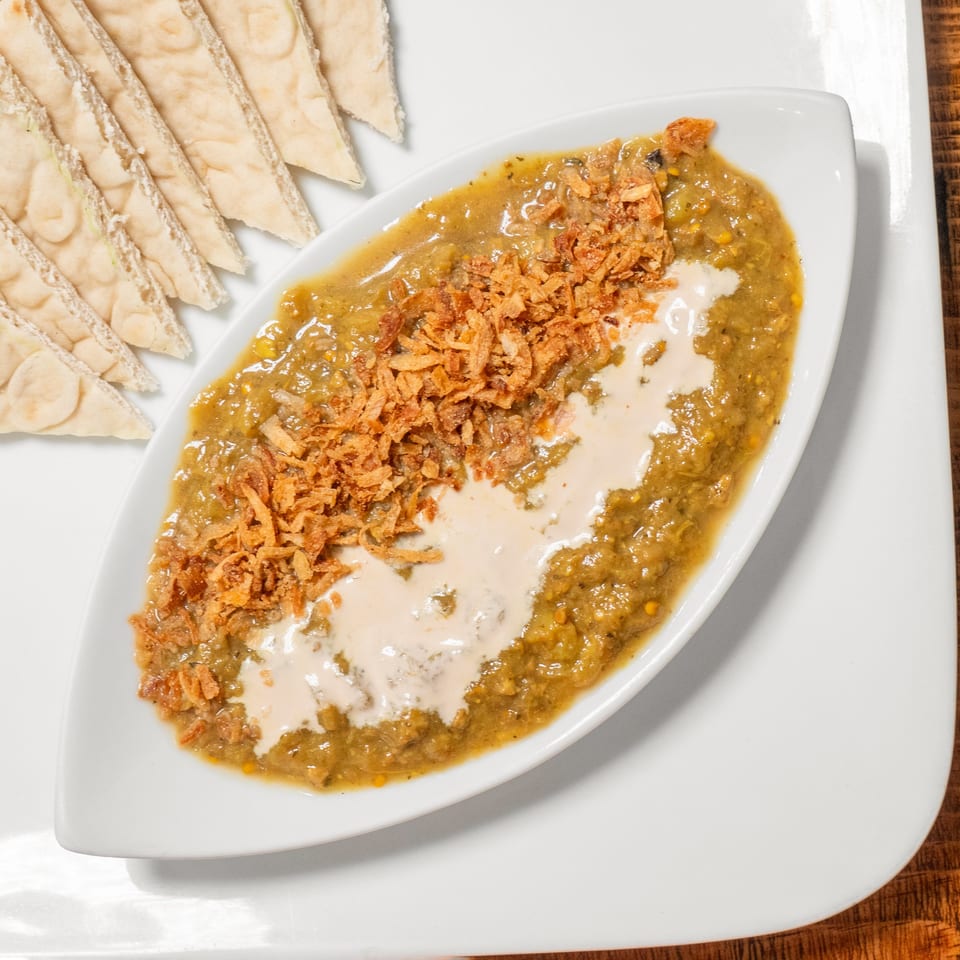 A white boat-shaped dish of green dip topped with fried onions and cream, served with pita bread.