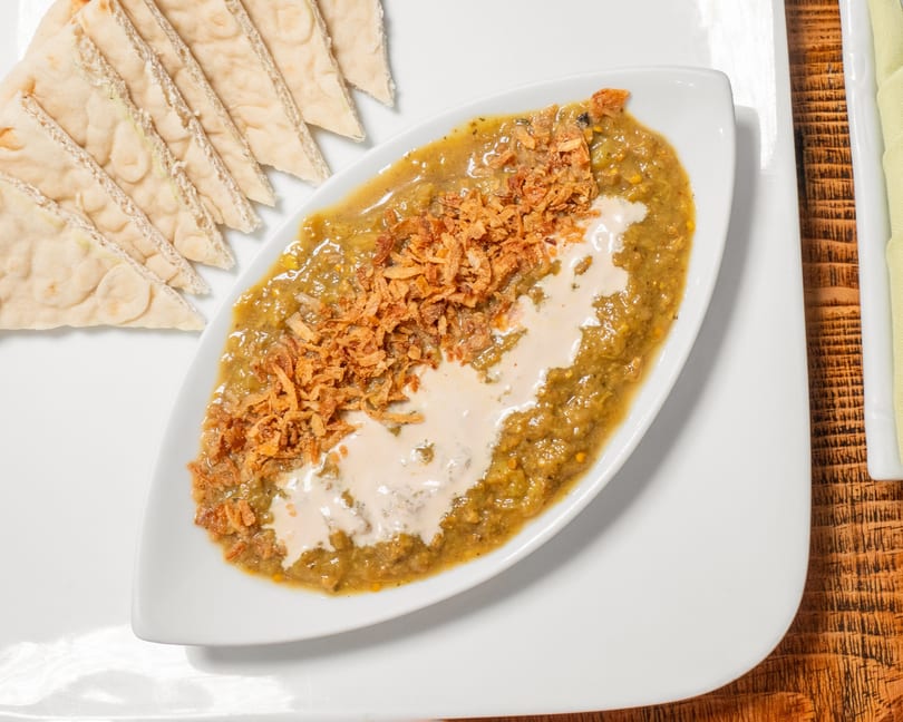 A white boat-shaped dish of green dip topped with fried onions and cream, served with pita bread.