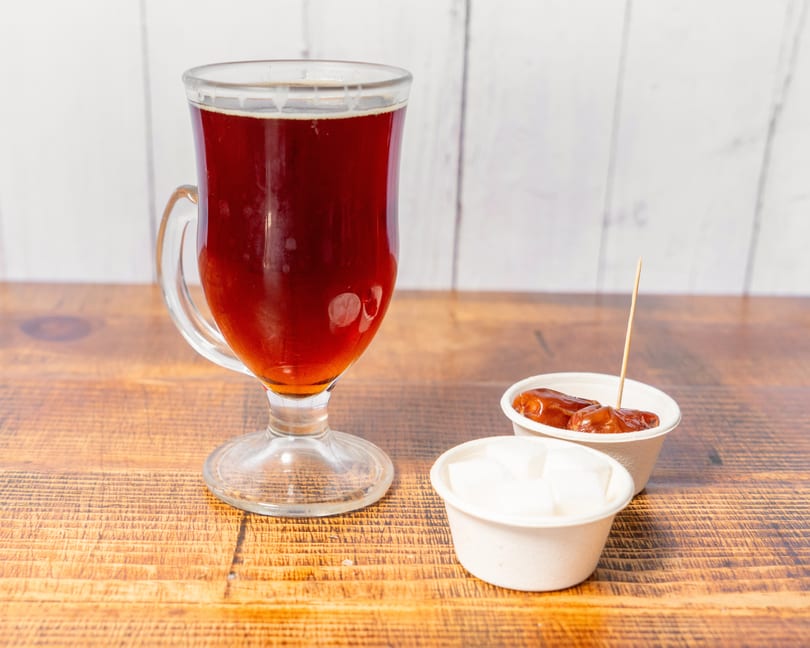 A glass of dark tea, with small cups of dates and sugar cubes on a wooden table.